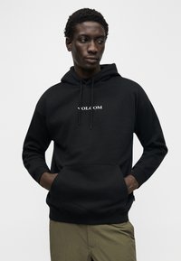 Sweat à capuche noir avec une poche avant, arborant un logo blanc "VOLCOM". Texture lisse, cordons de serrage ajustables et poignets côtelés.