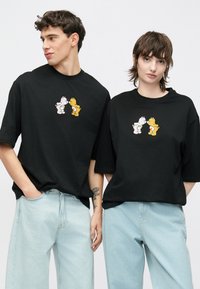 Μαύρα υπερμεγέθη t-shirts με γραφικά καρτούν αρκούδας σε λευκό και κίτρινο. Μαλακό ύφασμα με χαλαρή εφαρμογή, συνδυασμένα με ανοιχτό μπλε τζιν.