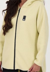 Gul fleecetröja med texturerad yta, hel dragkedja framtill och svart logotypmärke. Har huva och avslappnad passform.