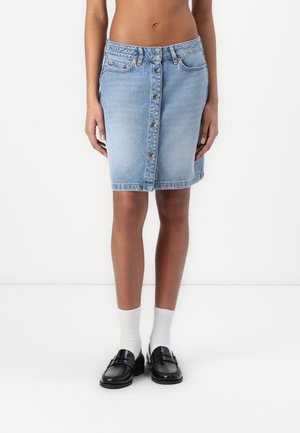 Lyseblå denimskjørt med knappelukking foran, til kneet, med to baklommer og en klassisk passform. Brukt med svarte sko og hvite strømper.