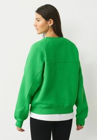 Zaļš kokvilnas džemperis ar pārāk lielu piegriezumu, ar ribotām aprocēm un apaļu kakla izgriezumu. Valkāts virs baltas krekla, ar vienkāršu teksturētu dizainu.