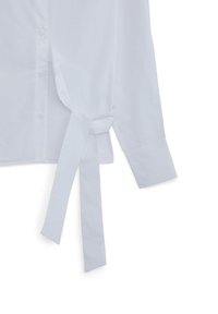 Camicia bianca a maniche lunghe con bottoni frontali e dettaglio di cintura in tessuto annodata di lato.