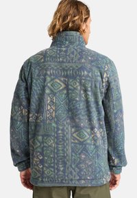 Fleece jacket in blue-green with geometric patterns in lighter shades. Features a high collar and side pockets. Soft texture and relaxed fit.