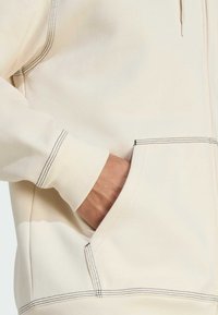 Sudadera de un tono beige claro con un bolsillo frontal y costuras en negro contrastantes. Cuenta con una capucha con cordón y puños de canalé para mayor comodidad.