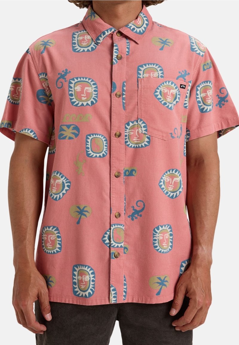 Chemise rose à manches courtes avec boutons, motifs tribaux bleus, beige et verts de visages, palmiers et lézards, portée sans être rentrée.