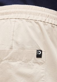 Κοντινή λήψη μπεζ ελαστικών σορτς με πίσω τσέπη και μαύρη ετικέτα λογότυπου στην άκρη της τσέπης.