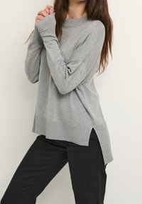 Maglione grigio lavorato a maglia con scollo e polsini a coste. Vestibilità leggermente oversize con spacchi laterali, abbinato a pantaloni neri. Texture morbida.