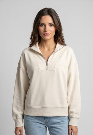 Mujer con cabello largo y marrón que lleva una sudadera cremosa con cremallera hasta un cuarto y vaqueros azul claro, de pie contra un fondo gris liso.