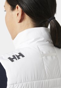 Mujer con cabello oscuro recogido en una coleta, vistiendo una chaqueta sin mangas blanca con el logo negro "HH" en el hombro.