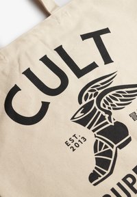 Bawełniana torba na zakupy w naturalnym kolorze z wyraźnym czarnym napisem "CULT" oraz grafiką buta z skrzydłami, założona w 2013 roku. Gładka tekstura.