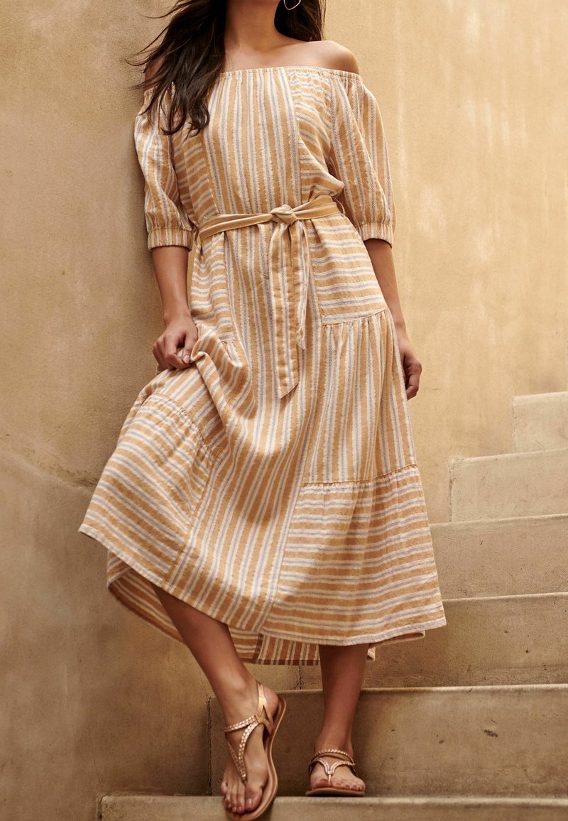 Femme portant une robe beige et blanche rayée, épaules dénudées, taille nouée, debout sur des escaliers en béton devant un mur beige texturé.