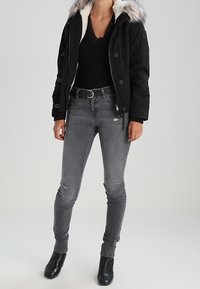Chaqueta negra con capucha y forro de pelaje sintético, camiseta negra, jeans grises desgastados y botines negros. El modelo está de pie en un entorno neutro.
