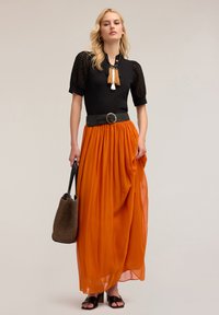 Maglietta nera con maniche texturizzate, gonna arancione fluida con pieghe, accentuata da una cintura nera con fibbia circolare e una borsa intrecciata marrone.