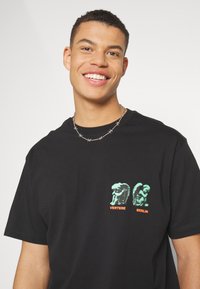 Black cotton T-shirt featuring green and orange graphic prints of sculptures with text "VERTERE" and "BERLIN." Round neckline and short sleeves.