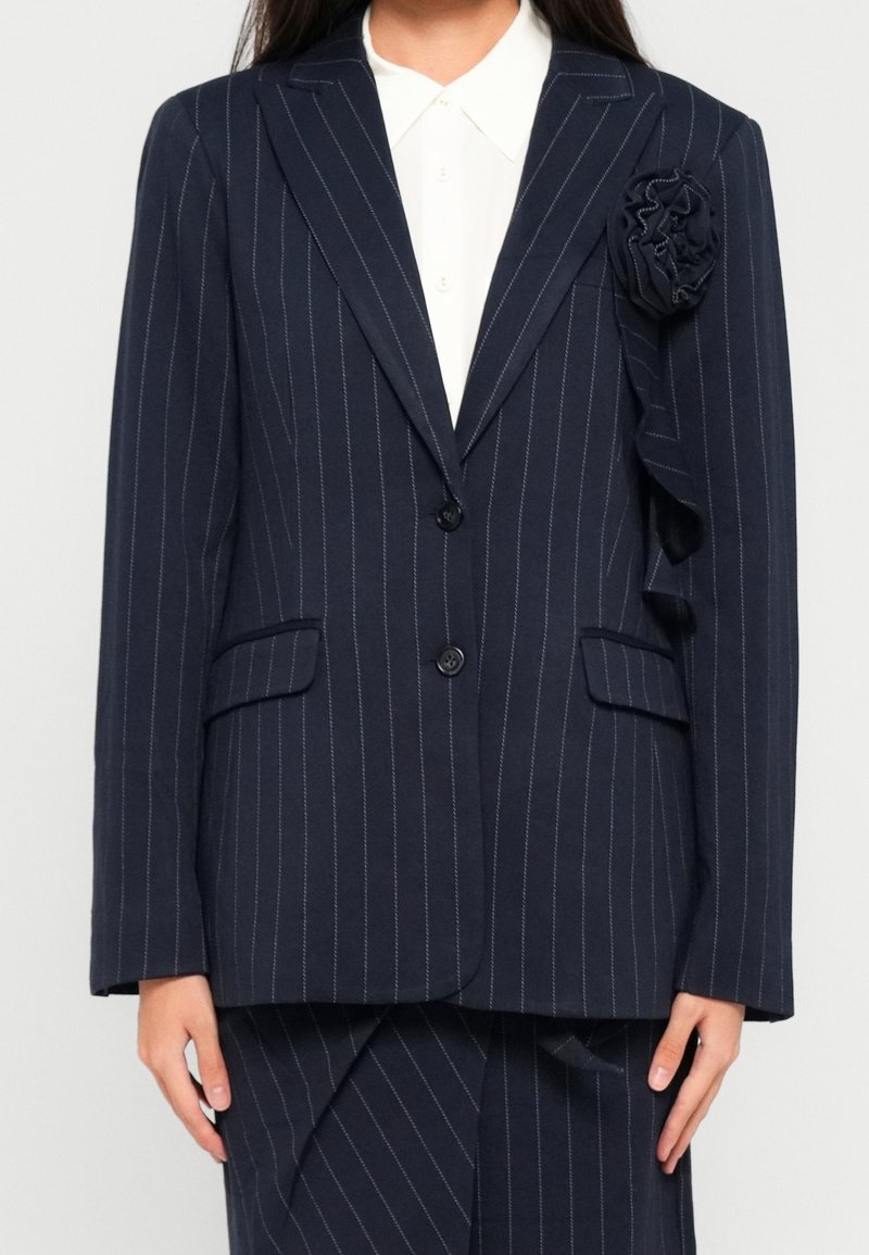 Veste bleu marine à fines rayures avec deux boutons, chemise blanche à col en dessous et détail d'une fleur en tissu sur l'épaule gauche.