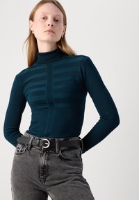 Teal tettskjorte med høy hals, i ribbestrikket tekstur og horisontale striper, kombinert med høyt belte svart jeans og et sirkulært spennebelte.