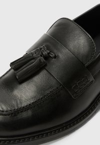 Mocassin en cuir noir avec une finition lisse, doté d'une sangle décorative et de glands. Bout rond et détails de couture subtils le long des bordures.