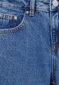 Gros plan sur un jean en denim bleu montrant la poche avant, la passants de ceinture, les rivets métalliques et les détails de couture.