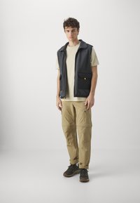 Gilet en polaire gris avec deux poches avant, porté sur un t-shirt beige. Pantalons cargo beiges avec poches zippées et chaussures de randonnée grises.