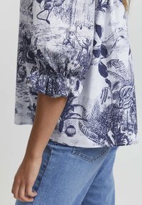 Top bianco con stampa botanica e fenicotteri blu navy, con maniche arricciate. Abbinato a jeans di denim blu chiaro.