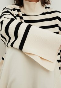 Pull en tricot rayé noir et crème avec un col montant et une texture côtelée, doté de manches larges et d'une coupe décontractée.