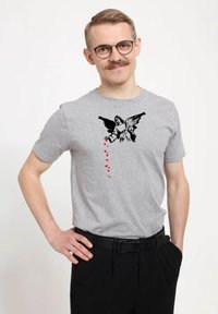 Usměvavý muž s brýlemi, který má na sobě šedé tričko s černým andělem, jenž nalévá červená srdce, která padají dolů, ruka na bocích.