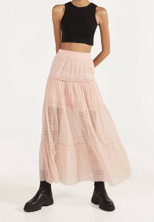 Kobieta ubrana w czarny bezrękawnik typu crop top, bladoróżową warstwowaną długą spódnicę oraz czarne masywne botki na kostkę na jednolitym białym tle.