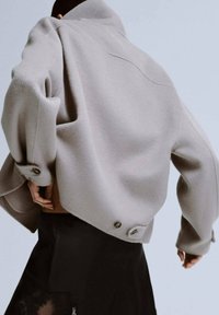 Lichtgrijze oversized jas gemaakt van zachte stof met een hoogstaande kraag, knoopdetails en een losse pasvorm. Draagt over een zwart kledingstuk.