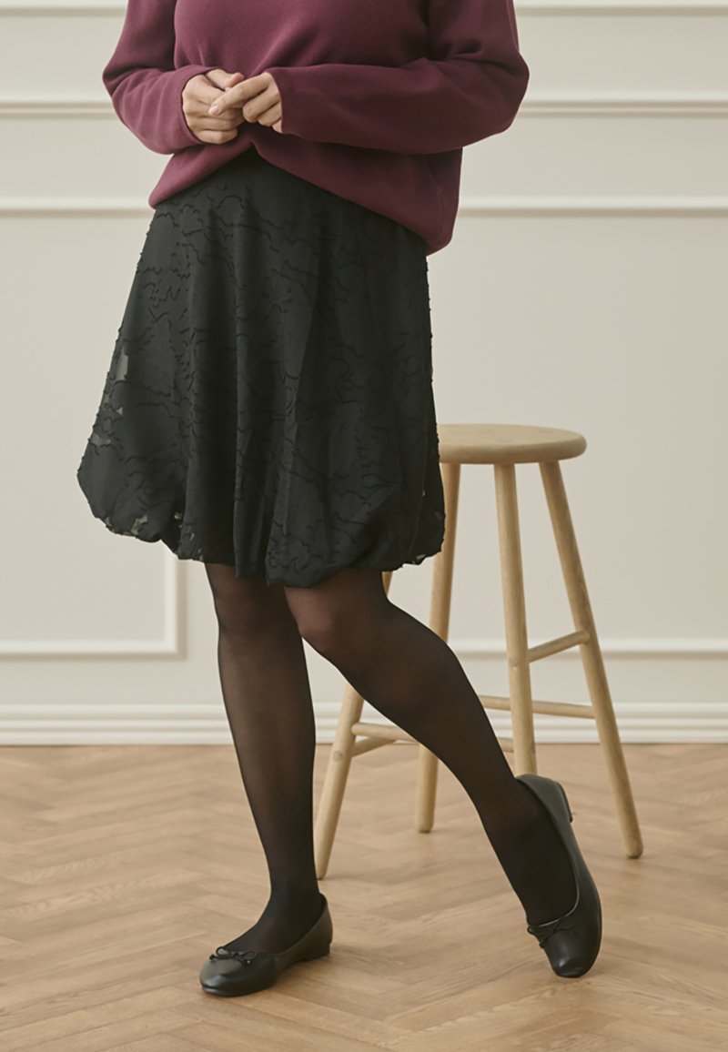 Črna krilo z teksturiranim cvetličnim vzorcem, kombinirano s črnimi neprosojnimi nogavicami in ravnimi čevlji z mašno. Nad tem bordo pulover.