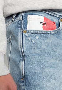 Jeans in denim azzurro chiaro con una texture consumata, dotati di una tasca bianca con dettagli rossi e neri e cuciture visibili.