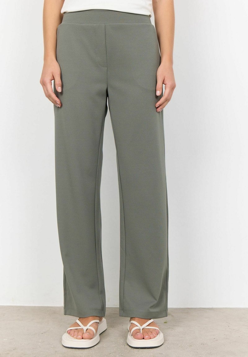Personne portant un pantalon large vert olive et des sandales blanches, debout contre un mur blanc uni et un sol en béton.