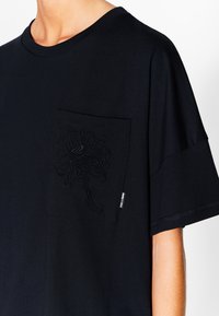 Camiseta de algodón negra con un bolsillo texturizado y un diseño floral bordado. Con mangas cortas y escote redondo.