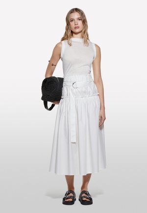 Haut sans manches blanc associé à une longue jupe blanche avec taille froncée, détail de ceinture et un sac tissé noir. Modèle portant des sandales noires.