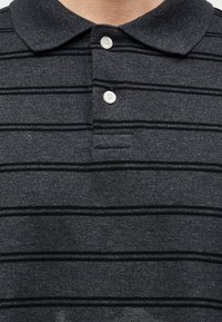 Σκούρο γκρι πόλο μπλουζάκι με μαύρες οριζόντιες ρίγες, δύο λευκά κουμπιά κλεισμένα κοντά στον γιακά στο πάνω μέρος του κορμού ενός ατόμου.