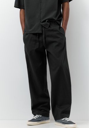 Személy laza fekete nadrágot és fekete, rövid ujjú, gombos inget visel, zsebébe tett kézzel áll, fehér háttér előtt.