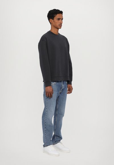 Un sweat-shirt crewneck gris foncé au fit décontracté, associé à un jean bleu clair et des baskets blanches, présenté sur un fond blanc.