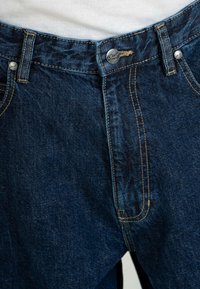 Jeans de mezclilla azul oscuro con corte recto, que presentan costuras amarillas, botones de metal y un diseño clásico de cinco bolsillos.