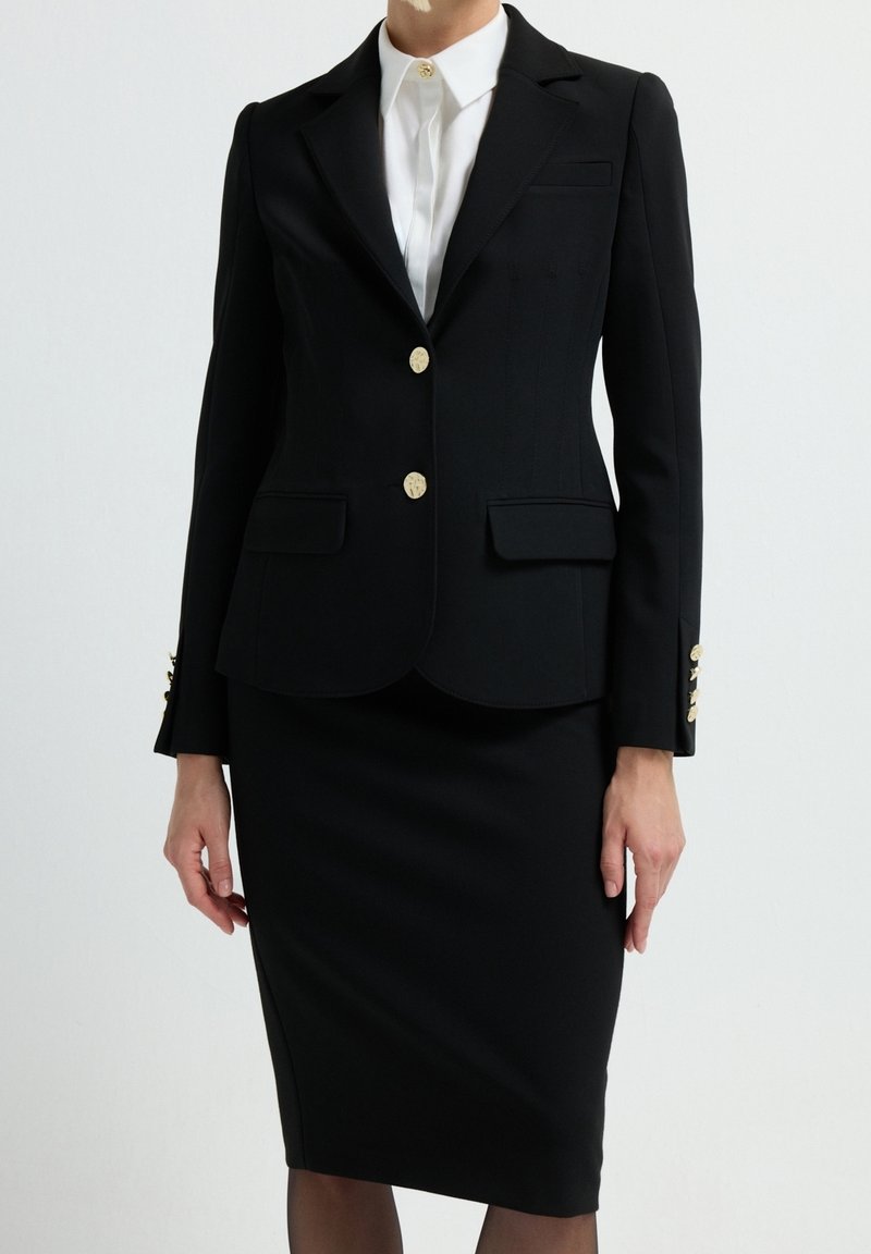 Femme portant un blazer noir cintré avec des boutons dorés et des poches à rabat sur une chemise blanche à col, associé à une jupe crayon noire.