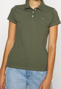 Olivgrön polotröja med klassisk krage, korta ärmar, treknappsstängning och en liten logotyp på bröstet. Tillverkad av bomullsblandat tyg.