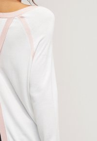 Donna che indossa un top bianco a maniche lunghe con bordo a costine rosa chiaro lungo il colletto e la cucitura della spalla.