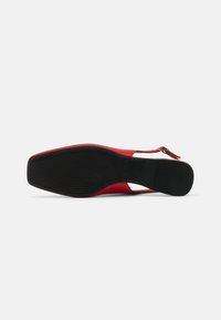 Escarpin rouge avec un talon ouvert, présentant une semelle en caoutchouc noir texturé, une tige lisse et un accent blanc contrastant près du talon.