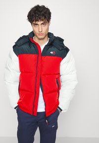 Chaqueta de plumas bicolor con un cuerpo rojo, mangas blancas y una capucha azul oscura, con cremallera frontal y parche de logo en el pecho.