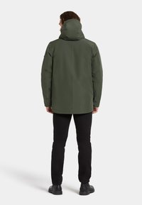 Vihreä vedenpitävä takki hupulla, pehmeä rakenne, rento istuvuus ja suora helma. Yhdistetty mustiin housuihin ja tukevasti kengät.