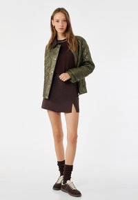 Veste matelassée olive avec un col doux, robe mini marron, chaussettes marron et baskets bicolores. La tenue comporte une fente sur le côté.