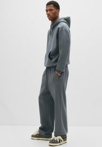 Ung mand står iført en matchende løs grå hættetrøje og joggingbukser med sort og hvide sneakers, hånd i lommen, neutral baggrund.