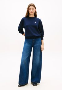 Σκούρα μπλε φούτερ με ριμπ μανσέτες και τελείωμα, με μικρό λογότυπο. Συνδυασμένο με φαρδιά σκουρόχρωμα τζιν παντελόνια και μαύρα παπούτσια.