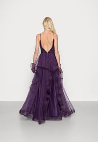 Fioletowa suknia wieczorowa z głębokim dekoltem na plecach, ozdobnym korpusem oraz lejącą się tiulową spódnicą z falbankami i delikatnymi ramiączkami.