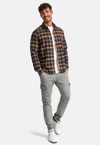 Karierte Hemd in Schwarz, Gelb und Pink über einem weißen T-Shirt, kombiniert mit hellgrauen Cargo-Hosen und weißen Turnschuhen. Person lächelt, Hände in den Taschen.