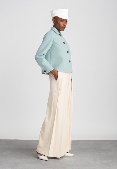 Fitted mint green jacket with black buttons, paired with high-waisted cream trousers and white fitted cap. Smooth texture, tailored design.