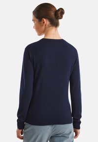 Mujer con cabello castaño recogido en un moño, vistiendo un suéter de manga larga azul marino y pantalones azul claro, mostrada de espaldas sobre un fondo blanco.